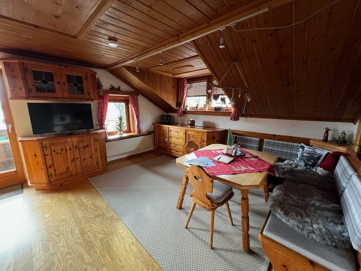 A cozy living area features warm wooden paneling and charming decor. A wooden table with a red tablecloth and chairs is positioned near a window. A television is mounted on the wall, and a sofa is placed against the side, enhancing the inviting feel of the room.