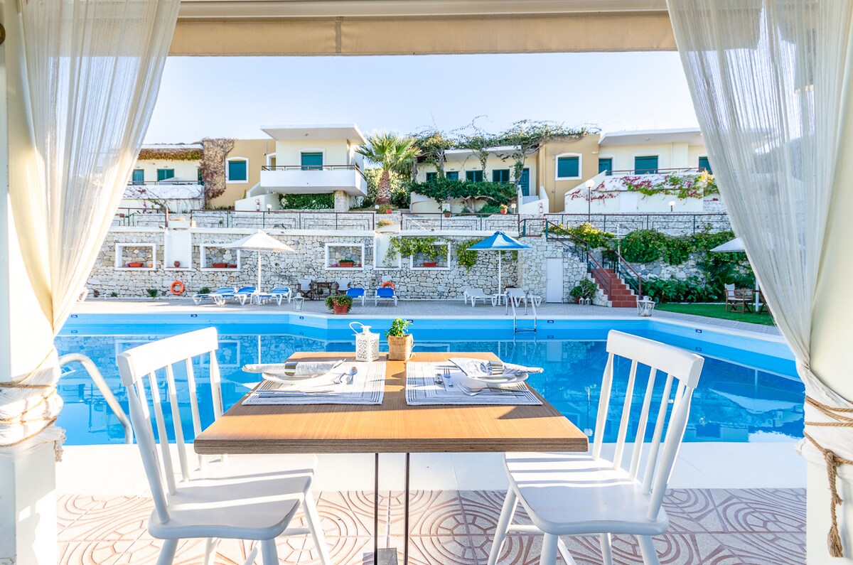 A dining table for two is set before a tranquil pool area, with a view of lounge chairs and umbrellas along the pool's edge. Lush greenery and terraced accommodations in the background contribute to a serene atmosphere.