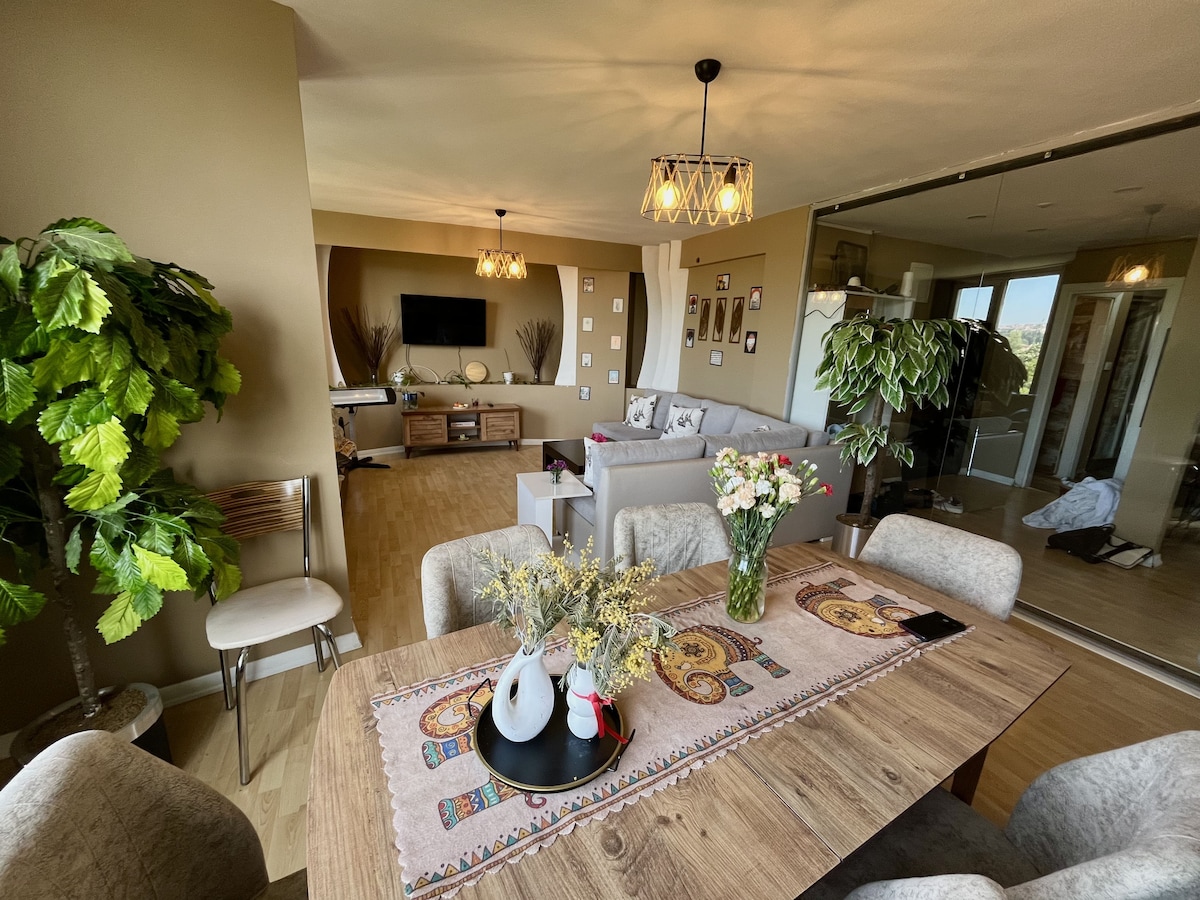 A dining area featuring a wooden table is surrounded by four upholstered chairs. A decorative runner is laid across the table, adorned with flowers and a vase. In the background, a cozy living area is visible with a sectional sofa and a TV on a console.