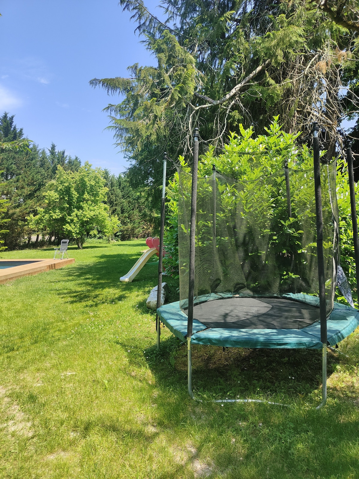 A spacious garden area is highlighted, featuring a trampoline surrounded by lush greenery. A slide is visible in the background, contributing to a family-friendly outdoor space. Sunlight filters through the trees, creating a bright and inviting atmosphere.