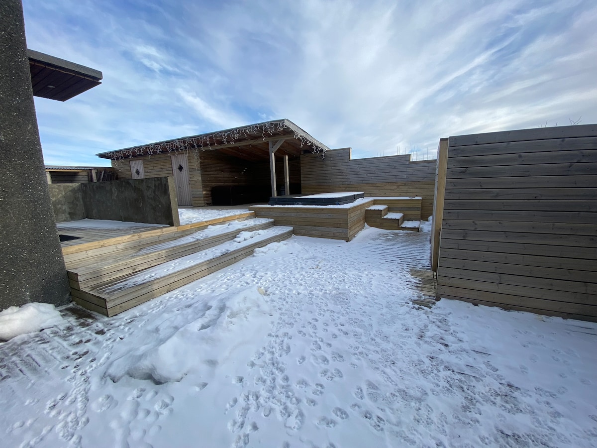 An outdoor area is visible, featuring wooden decking and steps leading to a covered seating space. The ground is blanketed by snow, with a clear sky overhead. The design promotes an open and inviting layout, ideal for relaxation in a natural setting.