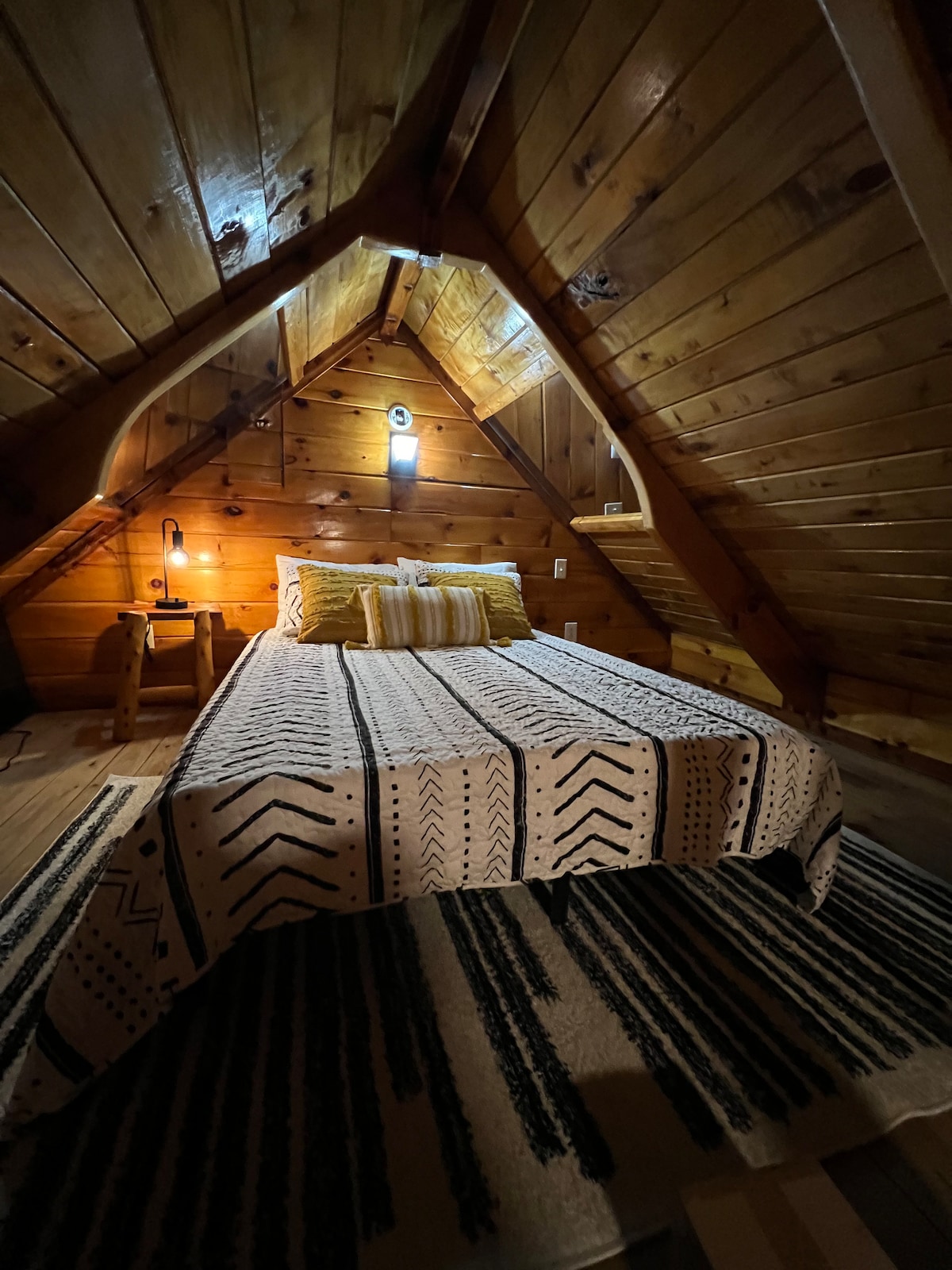 A cozy sleeping area is characterized by wooden walls and a sloped ceiling. A patterned blanket is draped over a spacious bed, complemented by soft lighting from a wall lamp and an accent table. A textured rug adds warmth to the wooden floor.