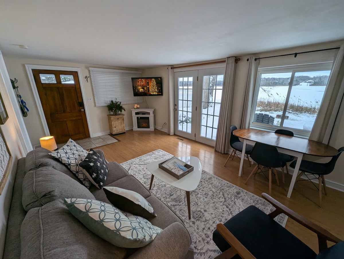 A spacious living area is highlighted by large windows offering a view of the tranquil outdoors. A comfortable gray sofa is positioned near a decorative fireplace, while a wooden dining table with chairs provides a cozy space for meals. Natural light fills the room.