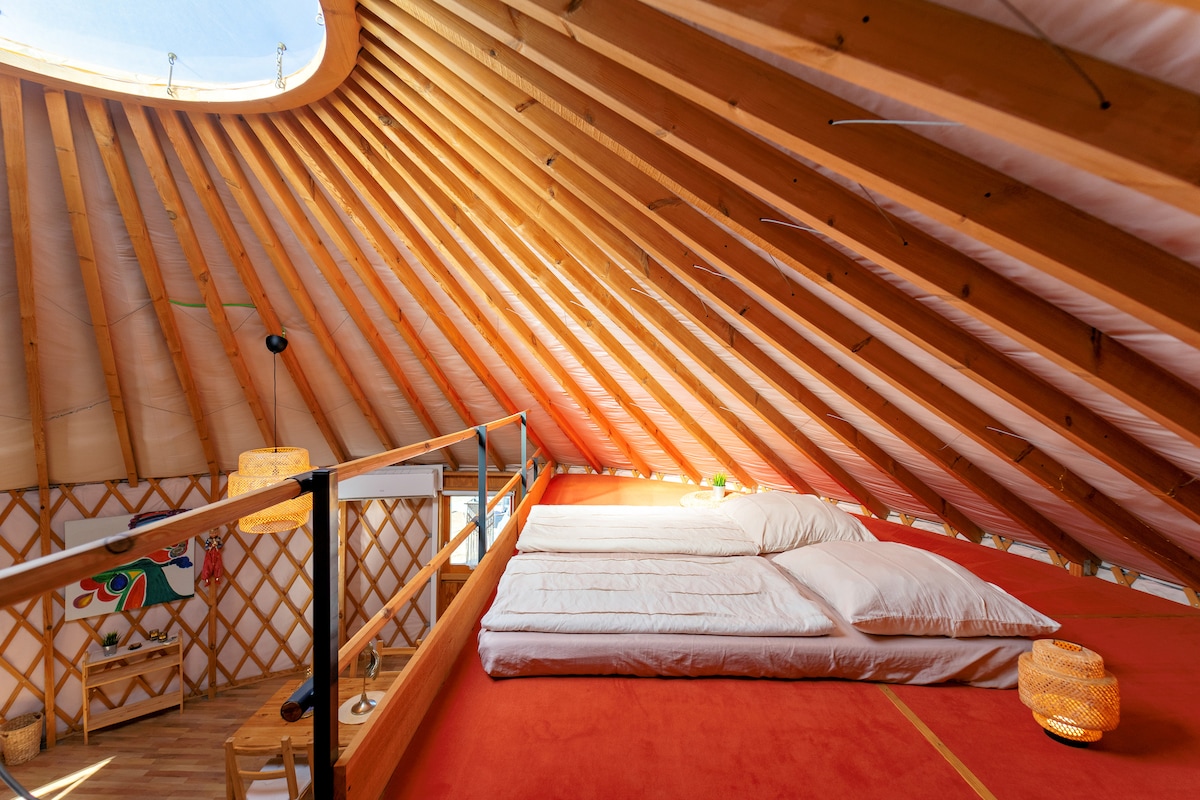 A spacious loft area features a large sleeping surface with soft bedding. Natural light enters through a circular skylight above, illuminating the wooden beams and cozy decor. A small light fixture is positioned nearby, providing additional warmth to the space.