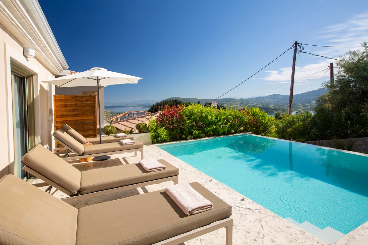 Lounge chairs are arranged around a swimming pool, offering a clear view of the surroundings. Umbrellas provide shade, and vibrant greenery frames the pool area. The horizon features a distant landscape under a clear blue sky, enhancing the serene outdoor atmosphere.