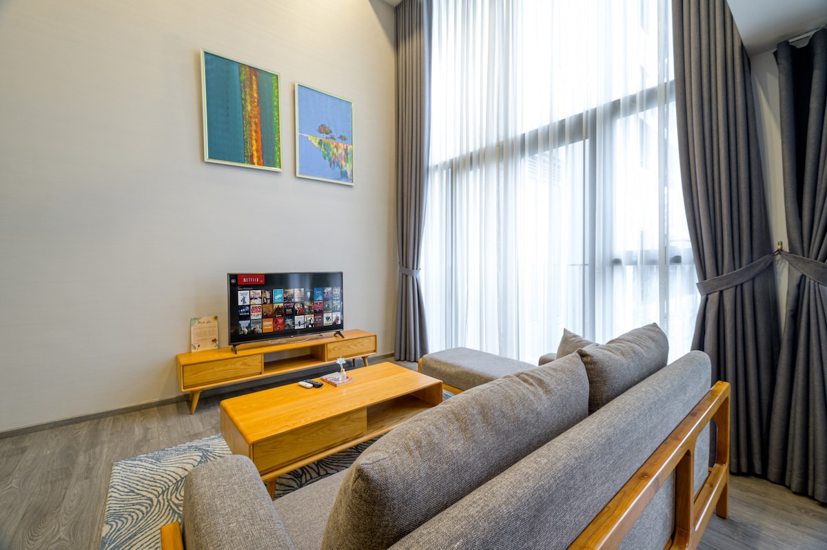 A spacious living area features a comfortable grey sofa, with a wooden coffee table in front. Two abstract paintings hang on the wall above the television, which displays a streaming service. Soft, natural light filters through large floor-to-ceiling windows, framed by sheer curtains.