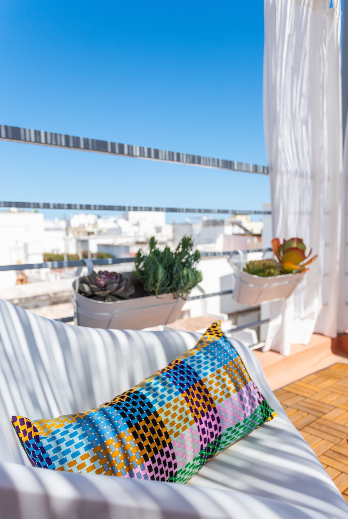 A cozy outdoor seating area is featured on the terrace, adorned with a colorful patterned pillow. Potted plants add greenery, while sheer curtains gently sway in the breeze against a bright blue sky.
