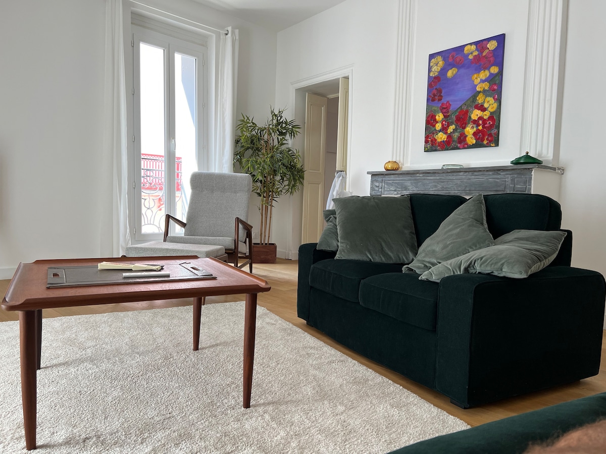 A spacious living area features a dark green sofa adorned with decorative cushions, accompanied by a stylish armchair. A wooden coffee table is positioned at the center, while a colorful floral painting decorates the wall. Natural light filters through the large windows.