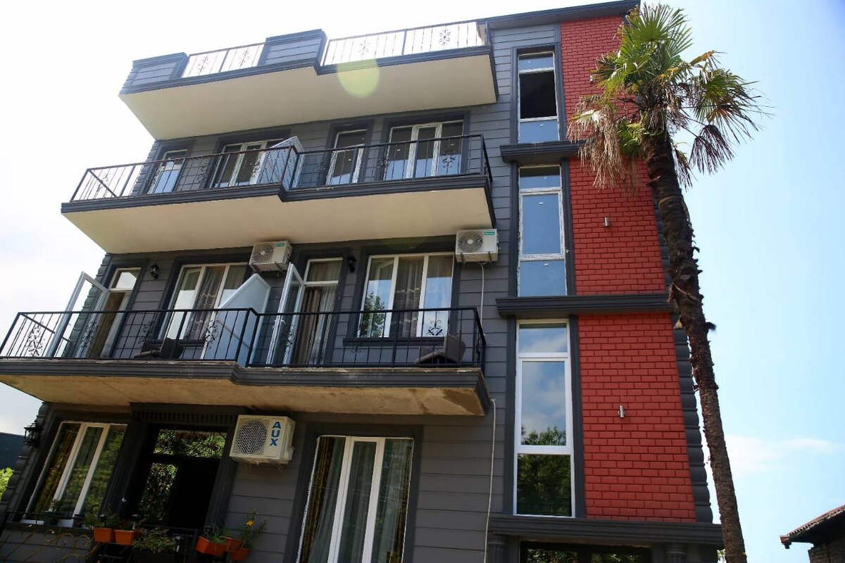 A modern three-story building showcases a mix of grey and red exterior walls. Balconies are visible on each floor, with several air conditioning units mounted on the façade. Lush palm trees and green landscaping are present at the base, enhancing the inviting appearance of the property.