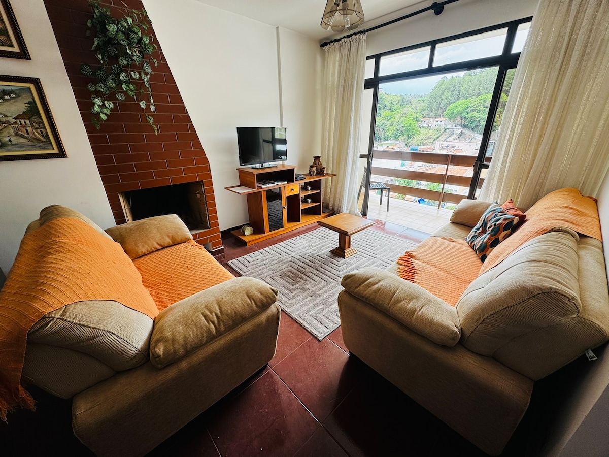 A spacious living area is highlighted by two comfortable sofas, arranged around a small coffee table. A television is positioned on a wooden stand against a wall. Large windows allow natural light to fill the room and offer views of the surrounding greenery.