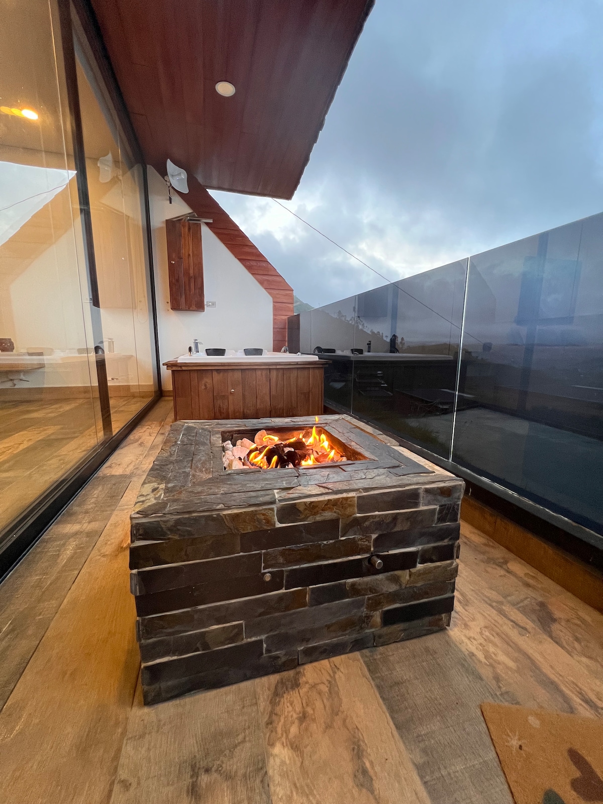 An outdoor space features a stone fire pit surrounded by smooth wooden flooring. Glass walls provide a panoramic view of the surroundings while soft evening light creates a welcoming atmosphere. The modern design includes a wood-paneled counter in the background.