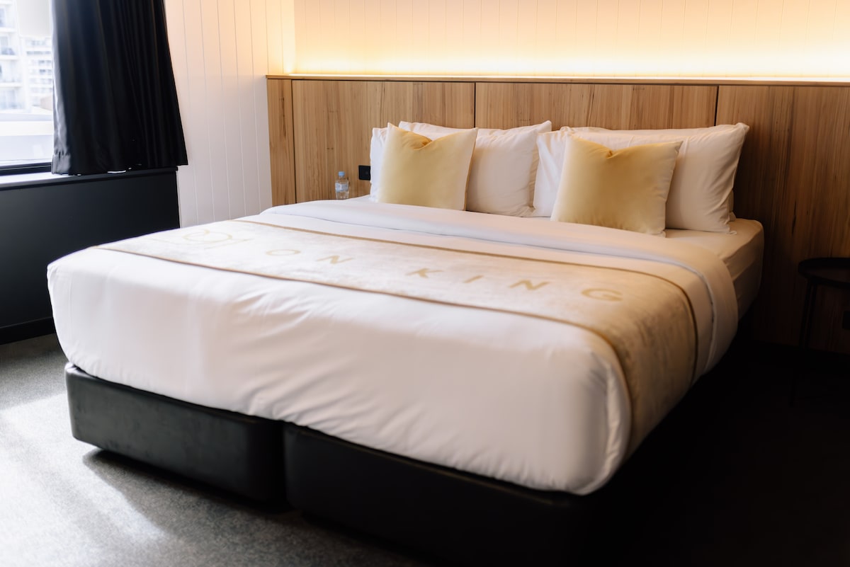 A spacious bed is positioned against a wooden accent wall, featuring three plush pillows. Soft lighting casts a warm glow above, creating a relaxing environment. A small black table is placed beside the bed, contributing to the room's clean design.