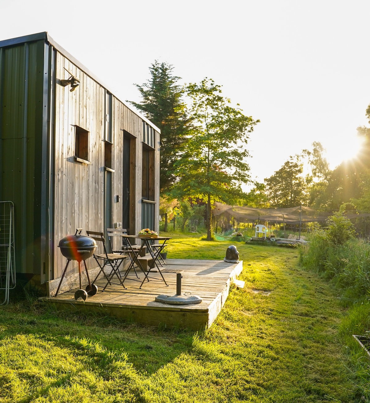 A wooden exterior of the refuge is illuminated by warm sunlight, showcasing a small terrace with two black chairs and a table. The surrounding greenery is lush, with a pathway leading into a natural landscape, creating a serene outdoor atmosphere.