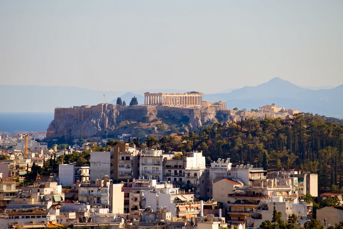 A panoramic view showcases the Acropolis, prominently positioned atop a hill, overlooking a densely populated cityscape. Surrounding buildings are visible, with layers of greenery in the foreground and distant mountains on the horizon, framed by a clear blue sky.