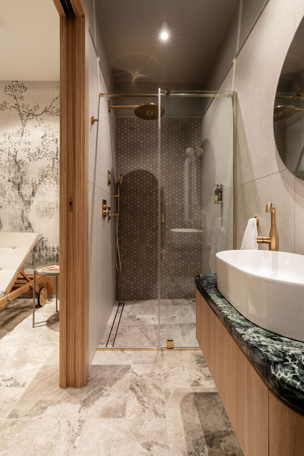 A modern bathroom features a walk-in shower enclosed by a glass panel. Elegant fixtures in gold tones are complemented by a round mirror above a vessel sink on a stone countertop. The textured wall and neutral tiles enhance the contemporary design.