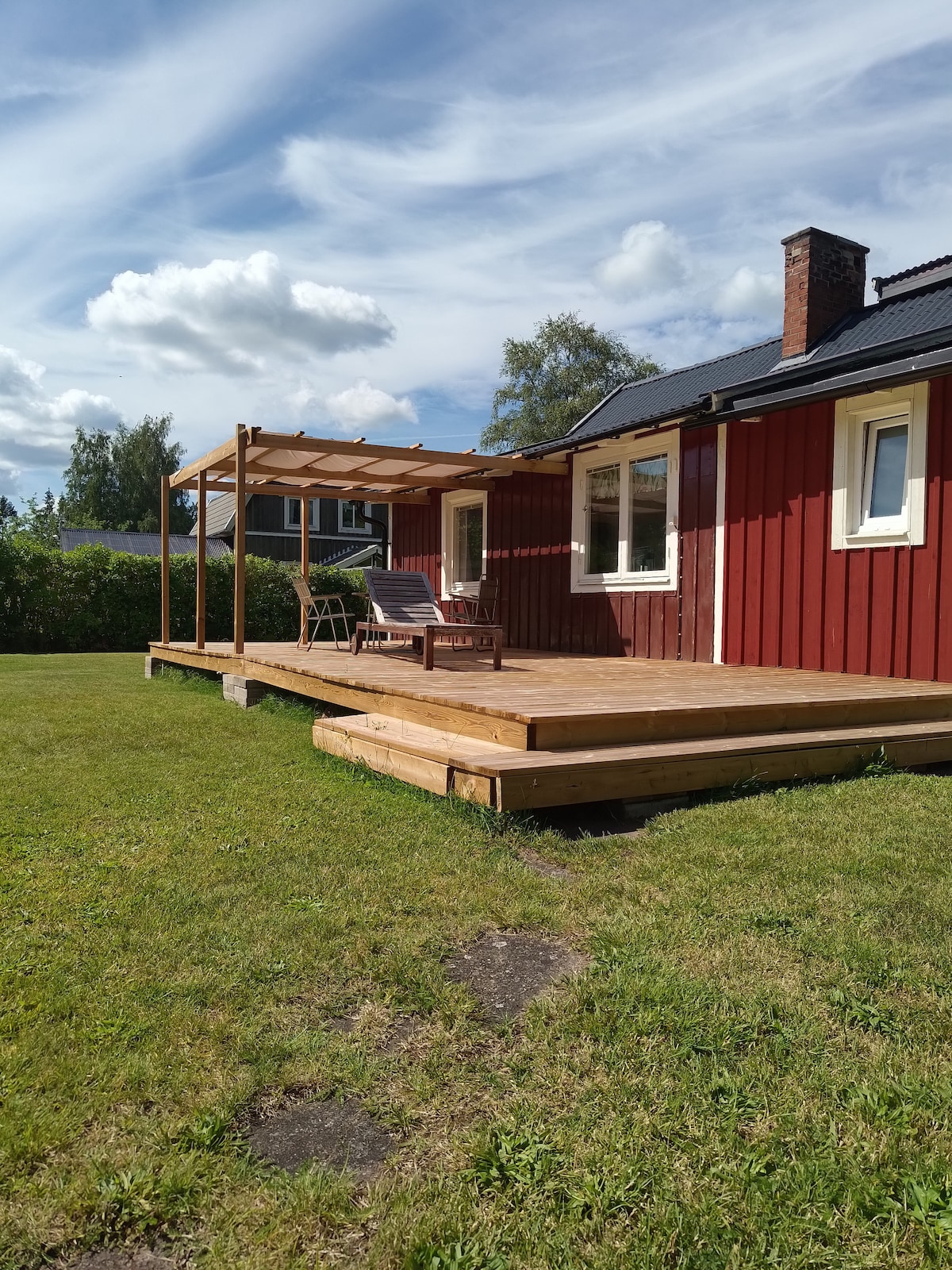 A wooden deck with a pergola is shown, featuring two chairs for relaxing. The red wooden exterior of the house contrasts with the green lawn, while fluffy clouds drift across a clear blue sky in the background.