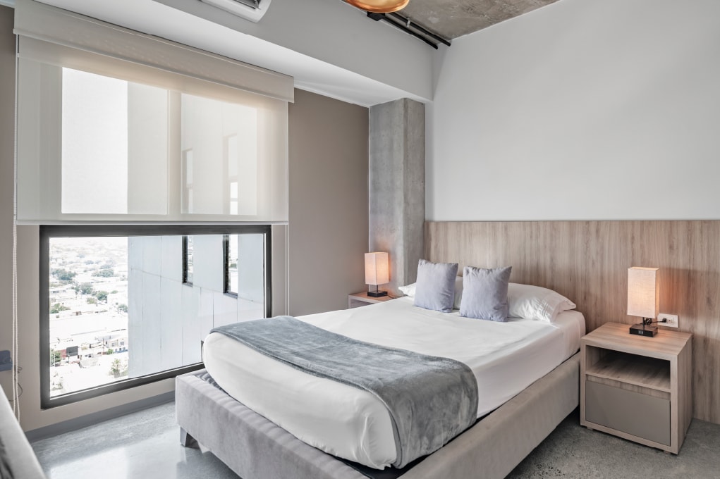 A tranquil bedroom is presented with a spacious bed, complemented by subtle grey and beige tones. Two bedside lamps provide gentle illumination, and large windows offer expansive city views, allowing natural light to fill the room.