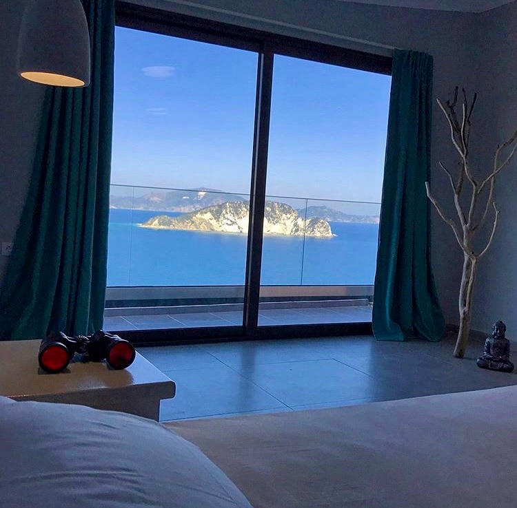 A spacious suite is showcased with large glass doors that open to a stunning sea view. Turquoise curtains frame the windows, while a small side table provides a place for binoculars, inviting exploration of the surrounding landscape. Natural light floods the room, highlighting the calm interior.