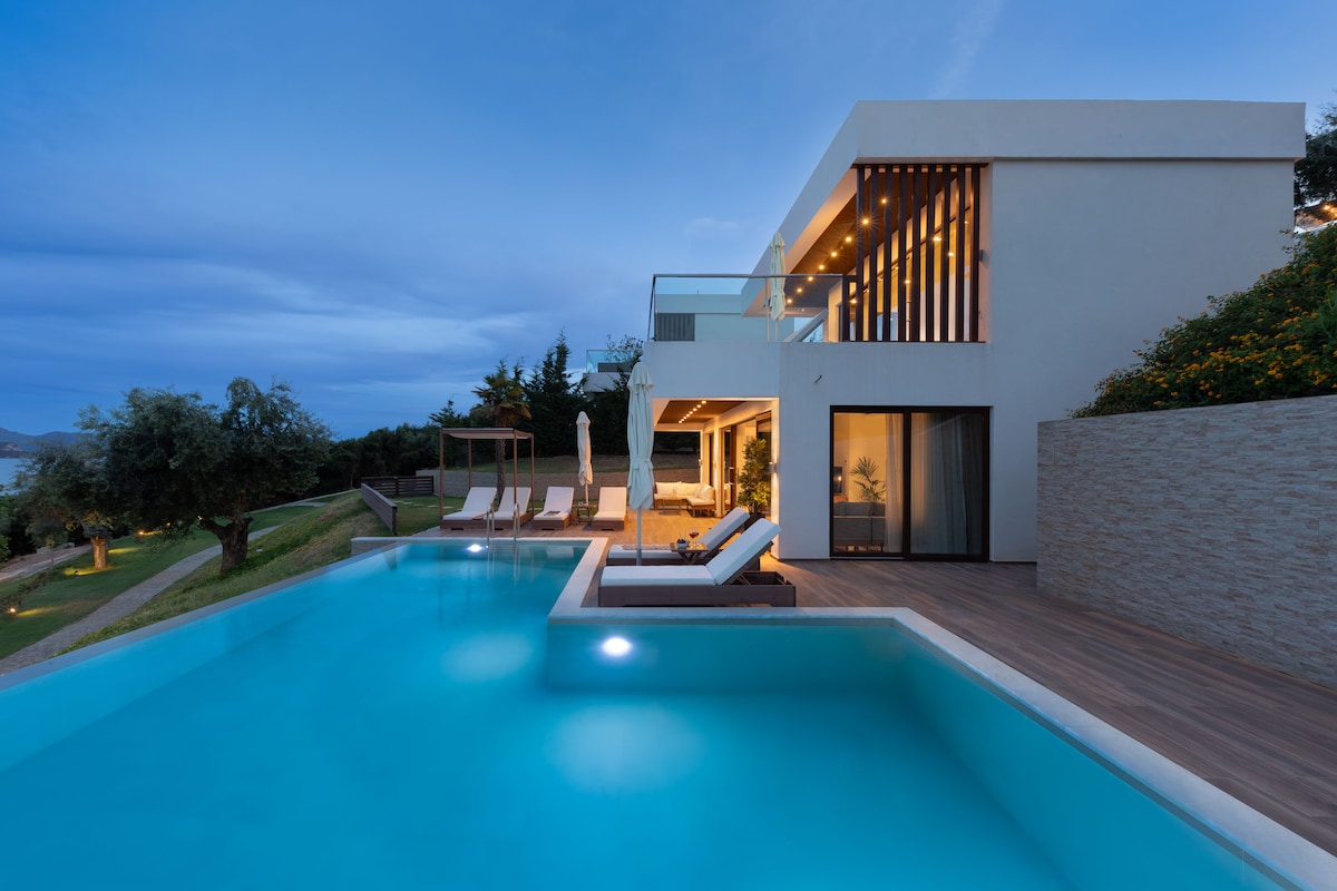 A modern villa is showcased beside a serene private pool, with a spacious deck featuring sun loungers and shaded areas. The architecture highlights clean lines and large glass doors, enhancing the connection between indoor and outdoor spaces. Natural surroundings provide a calming backdrop.
