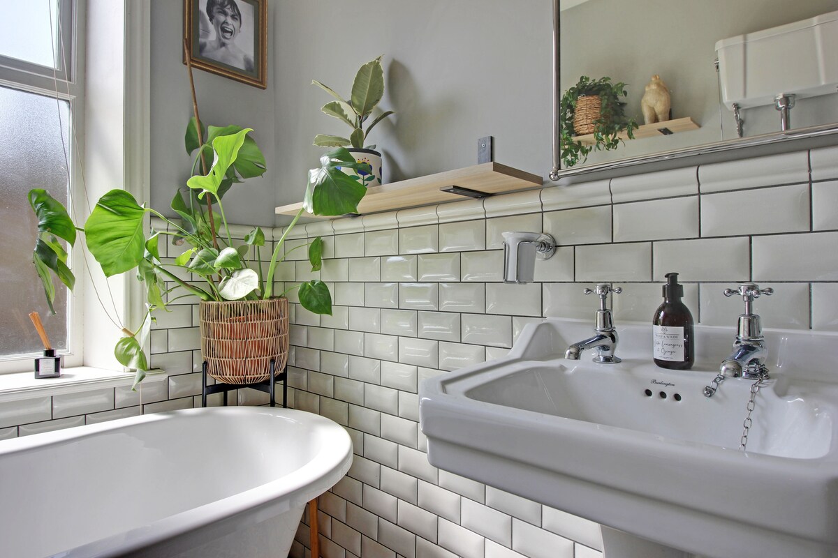 A modern bathroom features a roll-top bath beside a white, tiled wall. A plant in a woven basket adds a touch of greenery. A wall-mounted shelf displays decorative items, while a mirror reflects natural light from a nearby window. The sink is stylishly designed with vintage-style faucets.