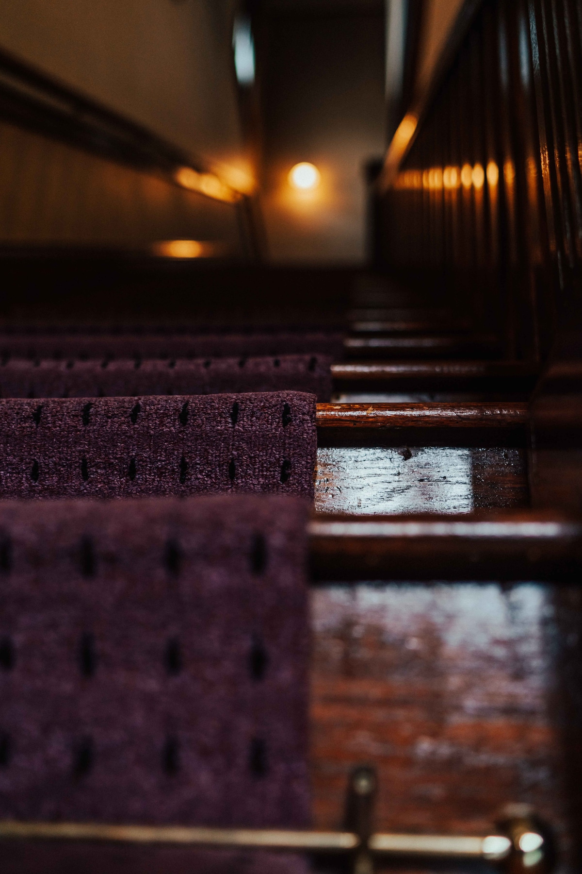 A close-up view of a staircase made from polished wood, featuring a deep purple carpet runner with a textured pattern. The warm glow of a wall-mounted light fixture illuminates the stairwell, creating a gentle ambiance.