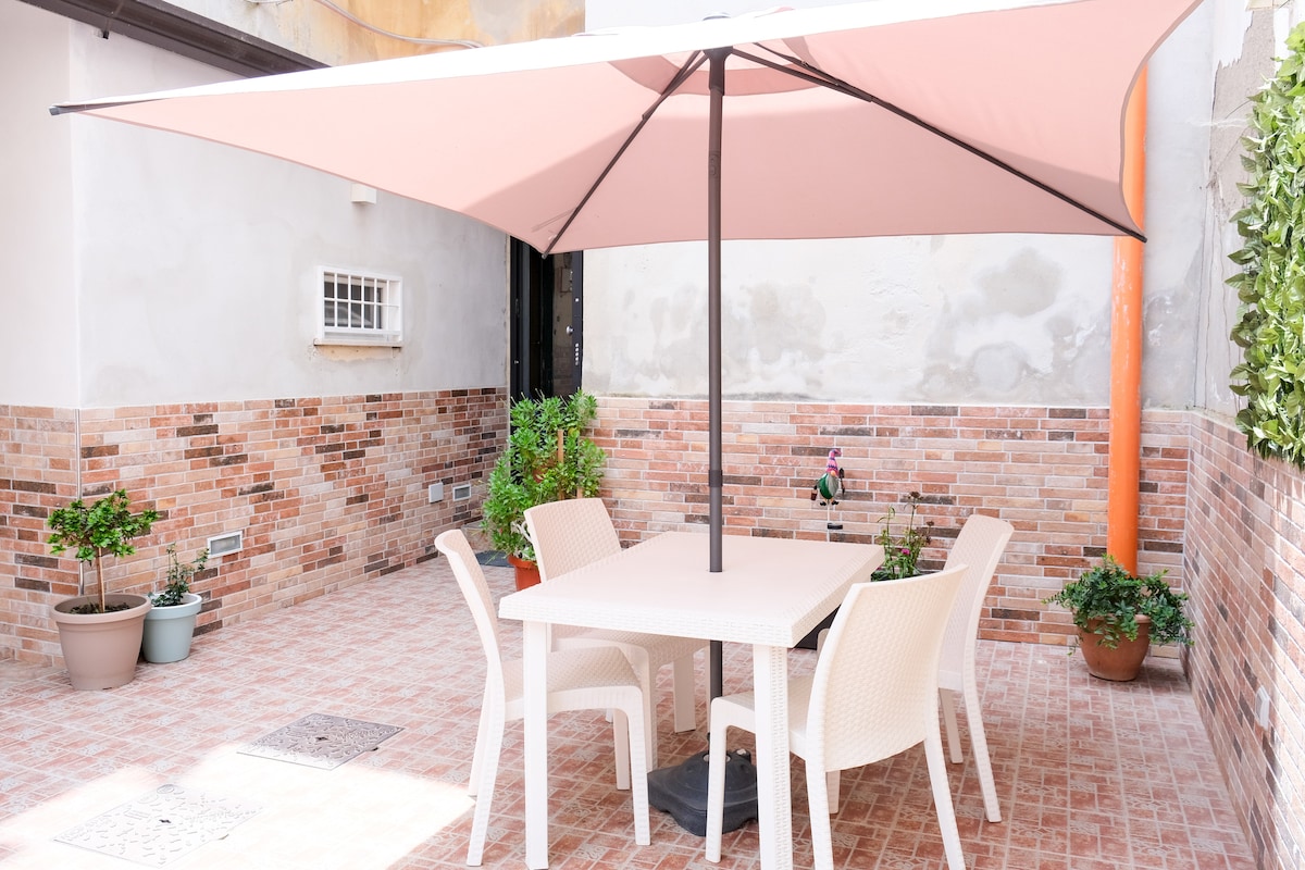 An outdoor seating area is presented with a square table surrounded by four chairs, all shaded by a large pink umbrella. Potted plants line the space, adding greenery to the brick-paved area enclosed by low walls.