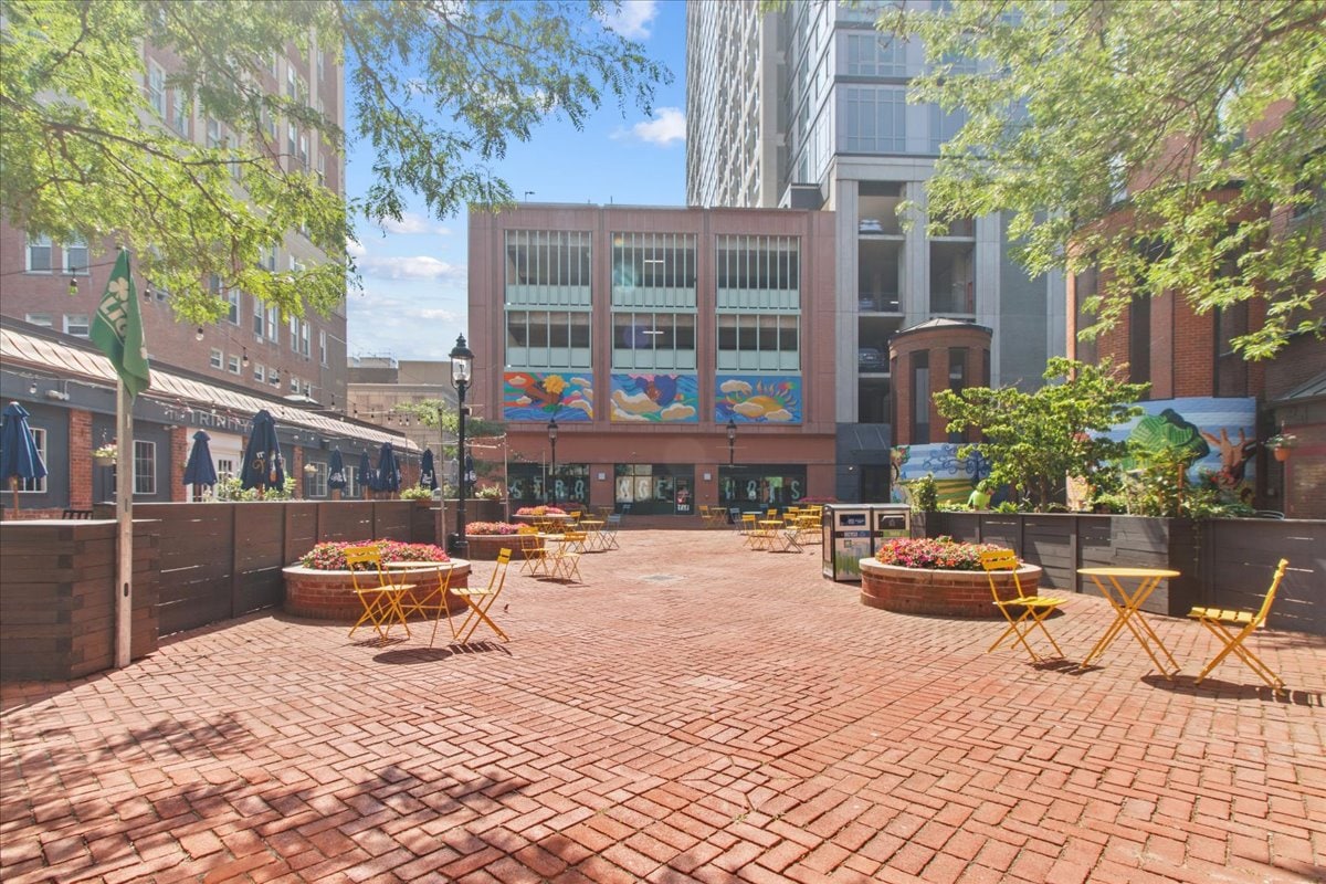 A spacious outdoor plaza is seen, featuring red brick pavement surrounded by greenery and wooden planters. Bright yellow bistro tables and chairs are arranged throughout the area, complemented by vibrant murals on the building facade, creating a welcoming atmosphere for relaxation.