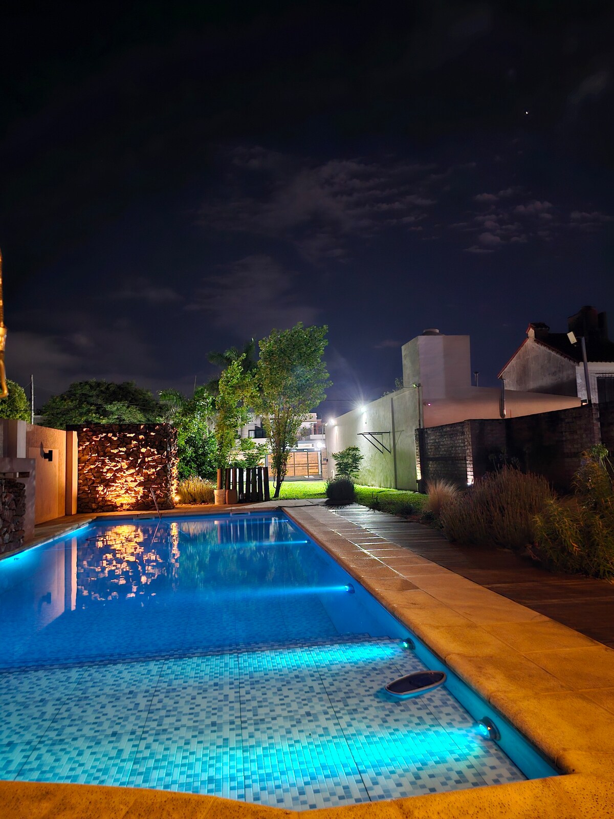 A well-lit outdoor area features a tranquil swimming pool reflecting gentle blue tones under the night sky. Illuminated trees and an adjacent stone wall enhance the serene ambiance, while soft lighting from nearby structures complements the setting.
