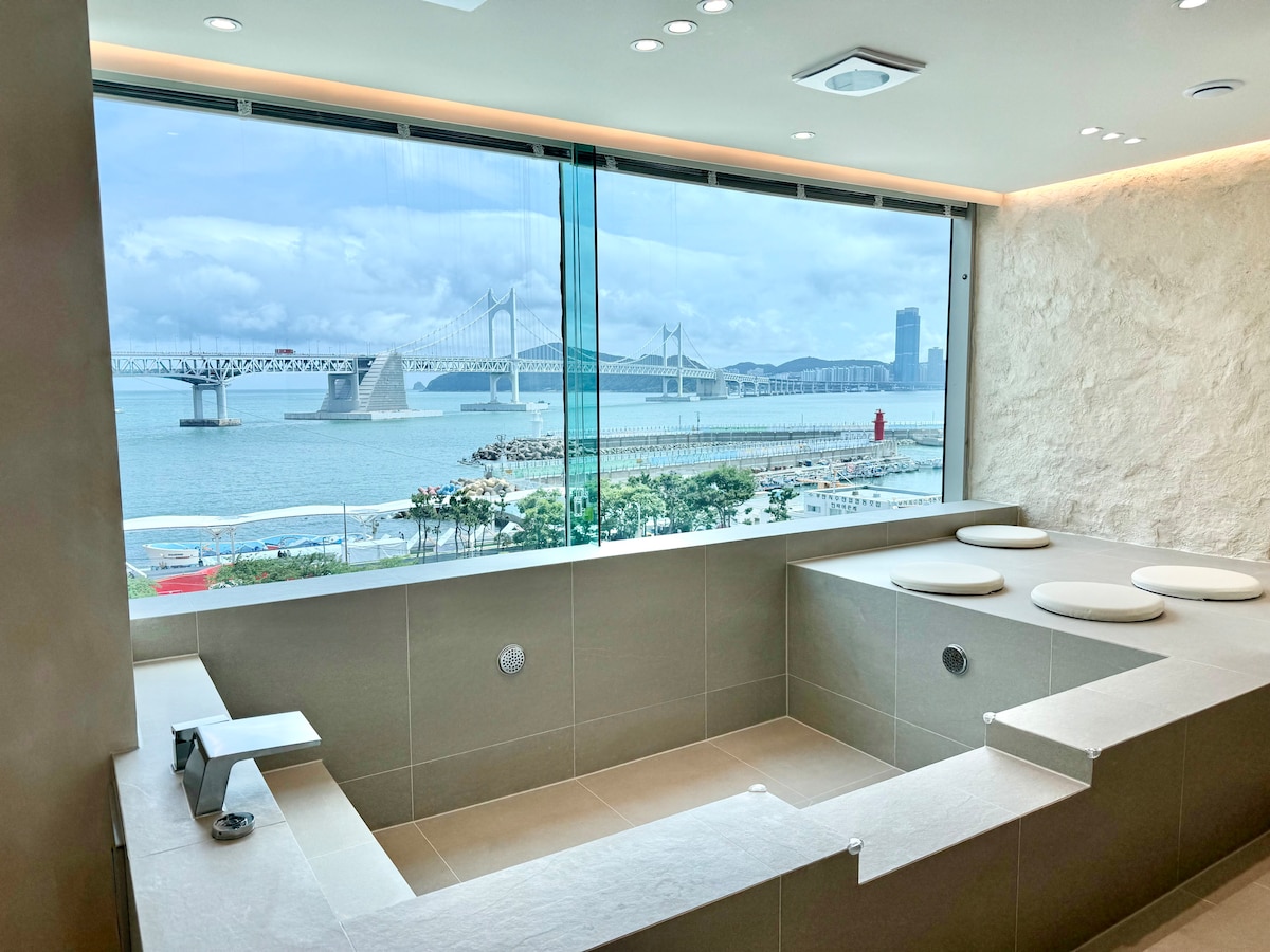 A spacious bathroom features a large soaking tub positioned by a panoramic window, offering views of the Gwangan Bridge and the coastline. Natural light fills the room, highlighting the modern tile design and minimalistic fixtures.