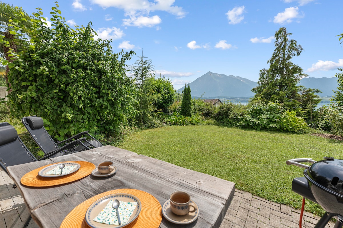An outdoor area features a wooden dining table set with plates and cups, surrounded by a lush garden. A gas grill and lounge chairs are included. In the background, a scenic view of mountains and clear skies can be seen, creating a tranquil setting for meals or relaxation.