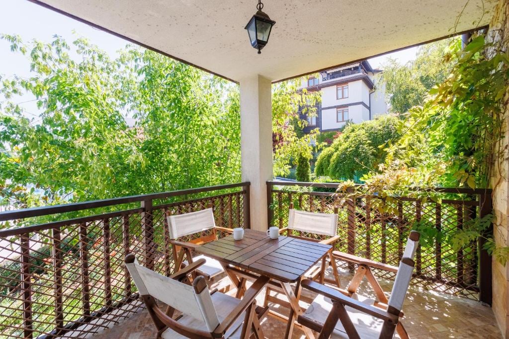 A furnished terrace features a wooden table surrounded by four white chairs, set against a backdrop of lush greenery. Natural light filters through the leaves, creating a refreshing outdoor space, suitable for enjoying meals or relaxation.