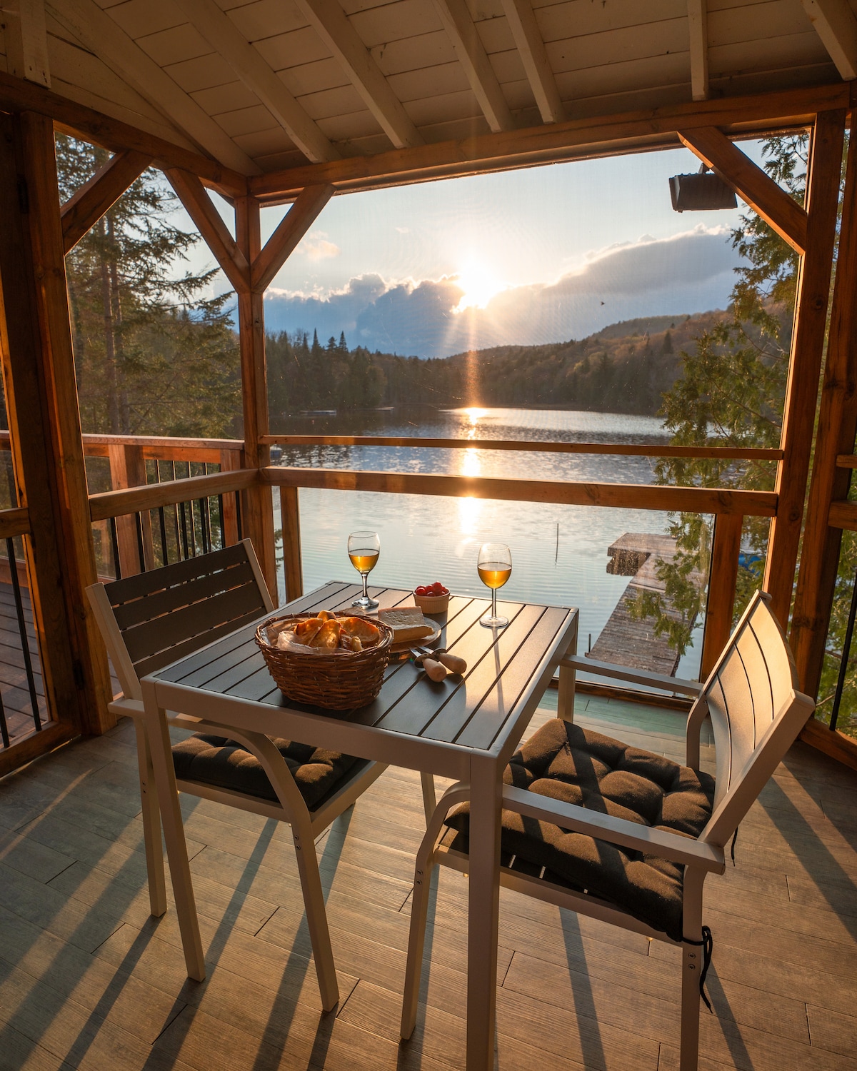 A serene outdoor seating area is observed, featuring a small table with a basket of pastries and two glasses. The view reveals a tranquil lake illuminated by the sunset, framed by trees, creating a peaceful atmosphere for relaxation.