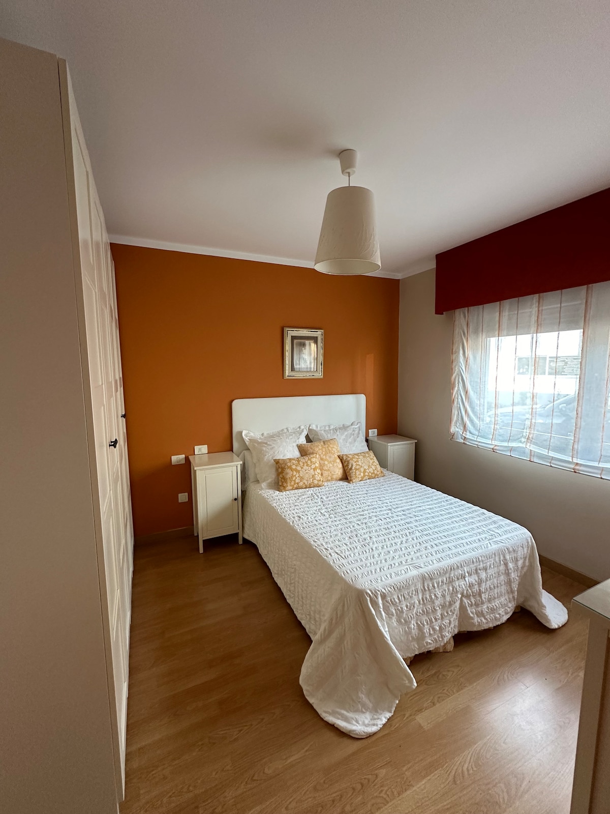 A bright bedroom features light wood flooring and an orange accent wall. A spacious bed is dressed in a white quilt, complemented by decorative pillows. Two nightstands flank the bed, each topped with soft lighting, while a large window provides natural light, adorned with sheer curtains.