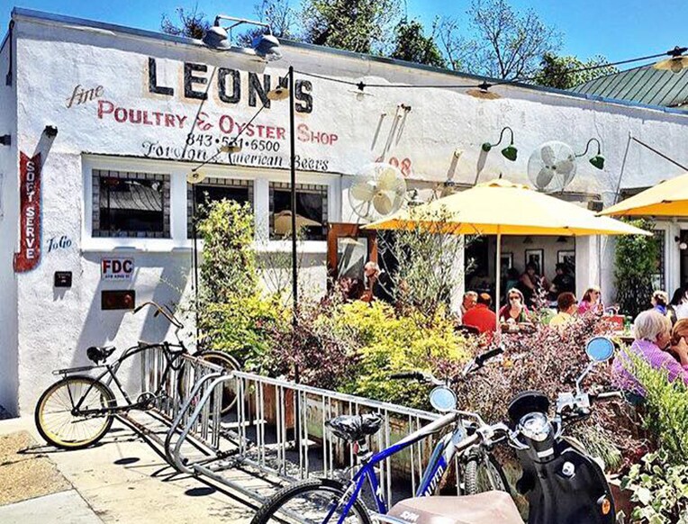 A charming eatery with a white exterior features outdoor seating shaded by yellow umbrellas. Bicycles are parked in front, and patrons are engaged at tables surrounded by vibrant greenery. The sign above displays the establishment's name and offerings, creating a welcoming atmosphere.