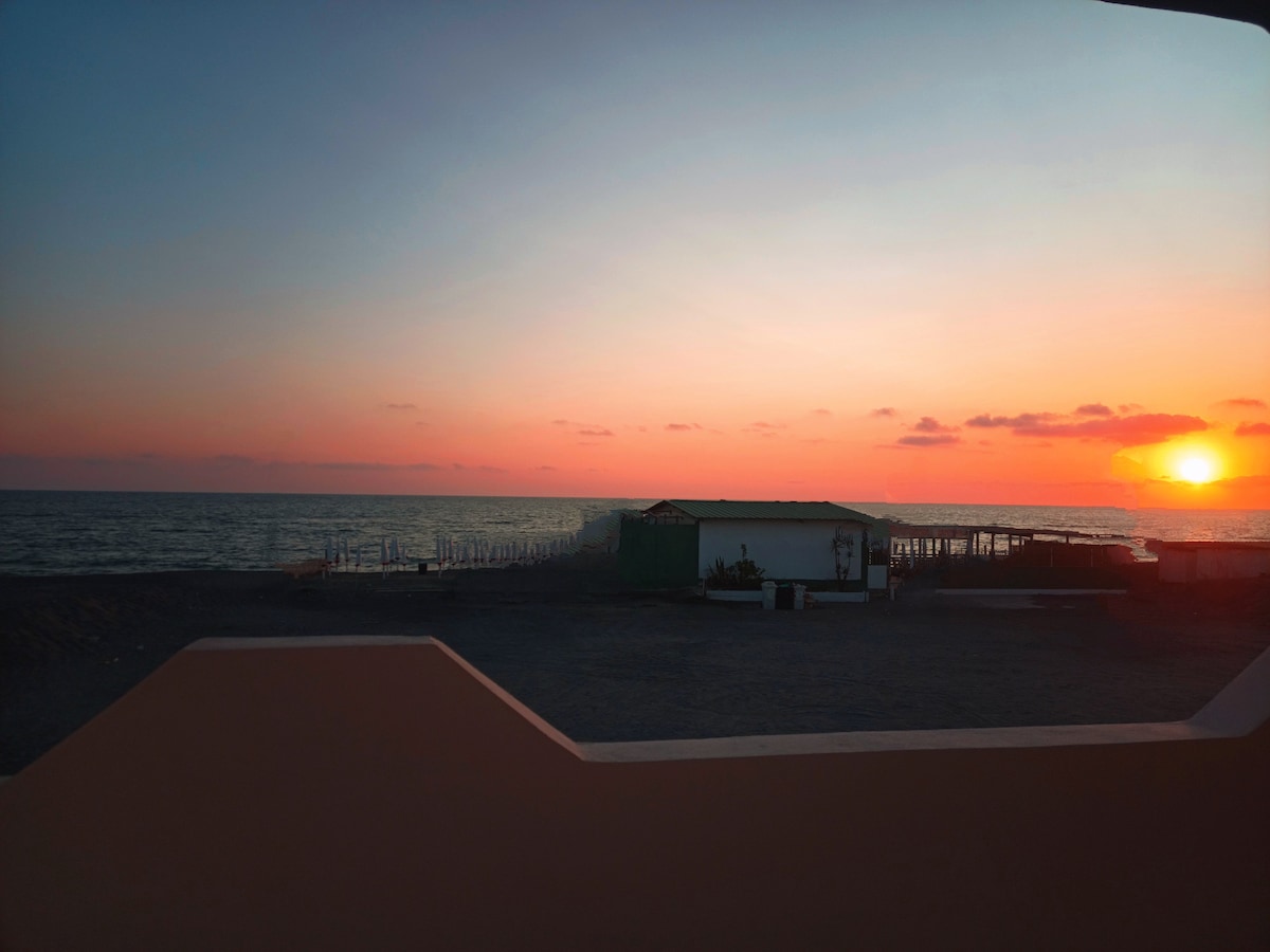The image captures a serene sunset over the Mediterranean Sea, with vibrant hues of orange and pink in the sky. A small beach hut is partially visible, along with the calm water stretching towards the horizon. The scene reflects a peaceful coastal atmosphere.