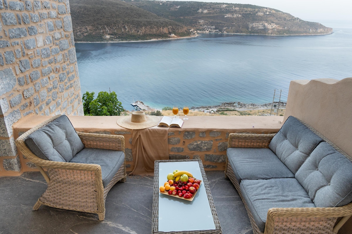 A cozy outdoor seating area features two woven sofas, complemented by a coffee table adorned with fresh fruits and a book. The backdrop includes panoramic sea views, enhancing the inviting atmosphere of the space.