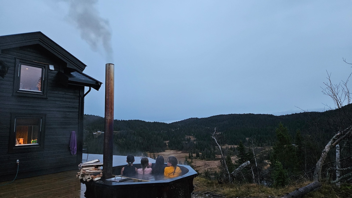 A wood-fired hot tub is set outside, with steam rising against a backdrop of rolling hills and trees. The adjacent black cabin features large windows and a cozy atmosphere. The scene suggests a tranquil outdoor experience for relaxation amidst nature.