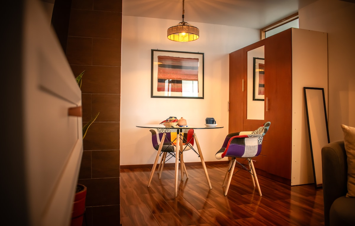 A modern dining area features a glass table surrounded by two colorful chairs. Warm wood flooring complements the space, while a pendant light hangs above. Framed artwork adorns the walls, and a partial room divider provides a sense of separation.