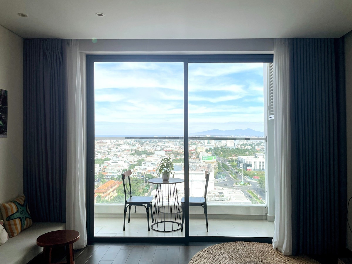 A large glass window offers a panoramic view of the city and coastline. A small round table is positioned in front, accompanied by two chairs. Natural light fills the space, highlighting the contemporary decor and soft color palette.