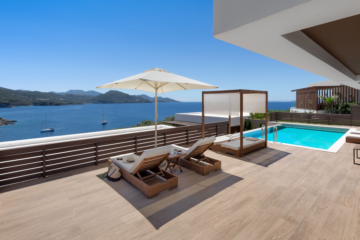 An outdoor terrace features comfortable lounge chairs and a sun umbrella, providing an ideal spot for relaxation. A private swimming pool glistens beside a cabana, with panoramic views of the Ionian Sea and surrounding mountains enhancing the serene atmosphere.