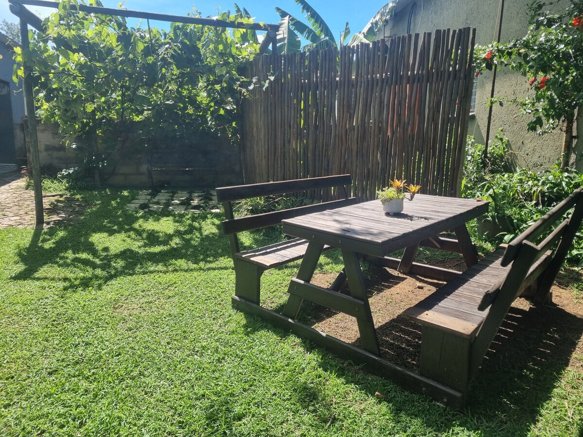 A wooden picnic table is surrounded by two benches in a lush green garden, offering a serene outdoor space. Bright flowers and climbing plants are seen nearby, with a bamboo fence partially enclosing the area, creating a sense of privacy and tranquility.