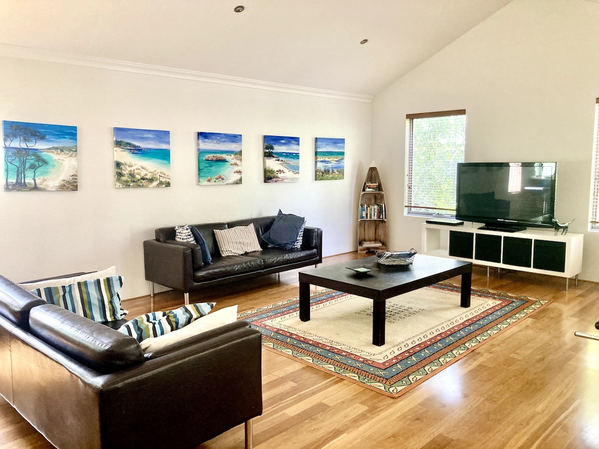 The open-plan living area features two leather sofas and a coffee table, with a large smart TV positioned on a white media unit. Colorful landscape artwork adorns the walls, contrasting with polished wooden floors and inviting natural light from the windows.