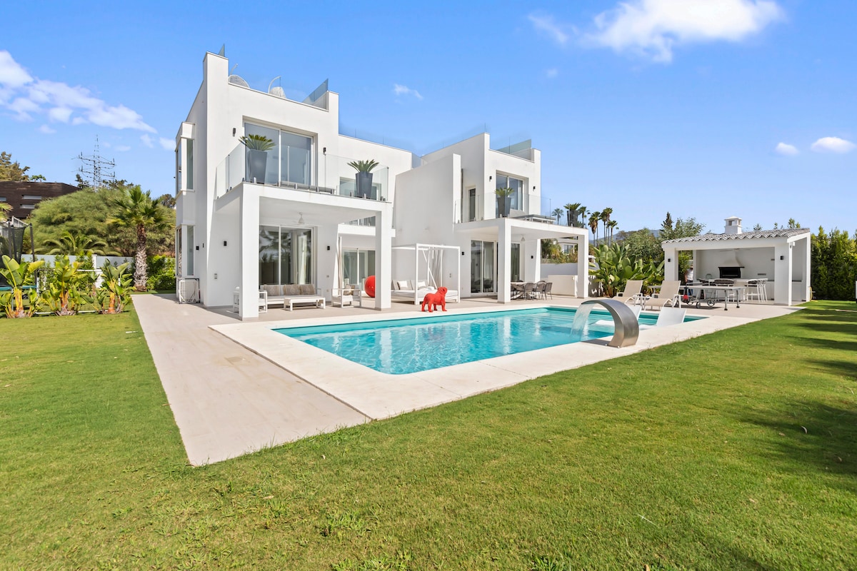 The villa features a modern architectural design, surrounded by lush green gardens. A spacious swimming pool reflects the clear sky, while multiple lounge chairs are positioned nearby for relaxation. The outdoor area is complemented by a vibrant red seating arrangement, enhancing the inviting atmosphere.