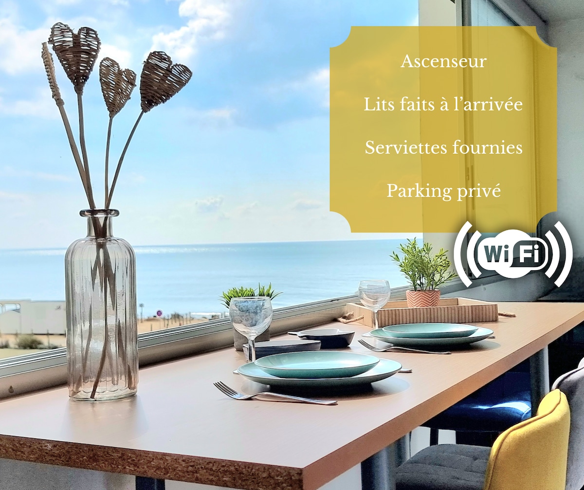 A dining table is set for breakfast with plates and glasses facing large windows that offer a seaside view. A decorative vase with dried flowers adds a touch of elegance. The tranquil sea can be seen in the background, enhancing the peaceful ambiance.