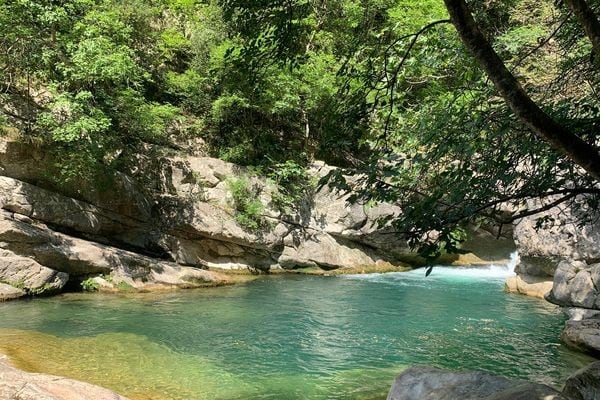 A serene natural pool is surrounded by rocky outcrops and lush greenery. The water reflects shades of blue and green, creating a calming atmosphere. The sunlight filters through the trees, casting gentle shadows on the water's surface. The scene invites relaxation and exploration of the natural beauty.