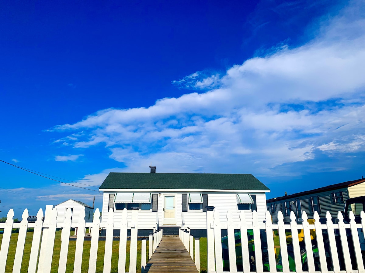 A quaint island cottage is framed by a white picket fence, with a wooden pathway leading up to the front door. The building features green awnings over windows and a clear blue sky adorned with soft clouds above.