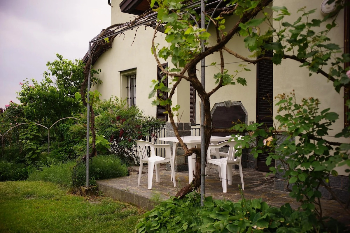 A shaded outdoor patio area is visible, featuring a white table surrounded by four plastic chairs. Green vines and lush bushes frame the space, while a stone pathway leads to a section of well-maintained grass, creating a serene outdoor setting.