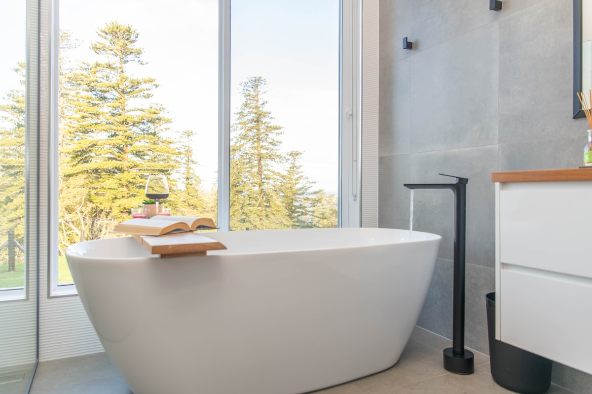 A modern bathroom features a freestanding soaking tub positioned near large windows that offer views of greenery outside. A wooden tray rests on the tub, holding a book and a glass. The minimalist design is complemented by a stylish black faucet and a sleek vanity.