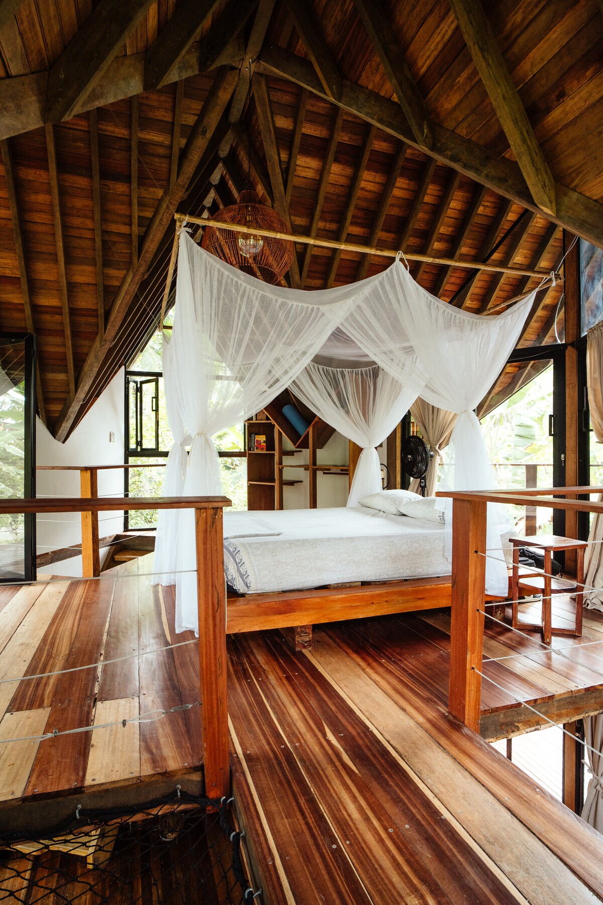 A spacious bedroom features a wooden floor with a natural finish and a high, slanted ceiling. A large bed is draped with light, sheer fabric, creating a serene sleeping area. Large windows allow natural light to fill the space and provide views of surrounding greenery.