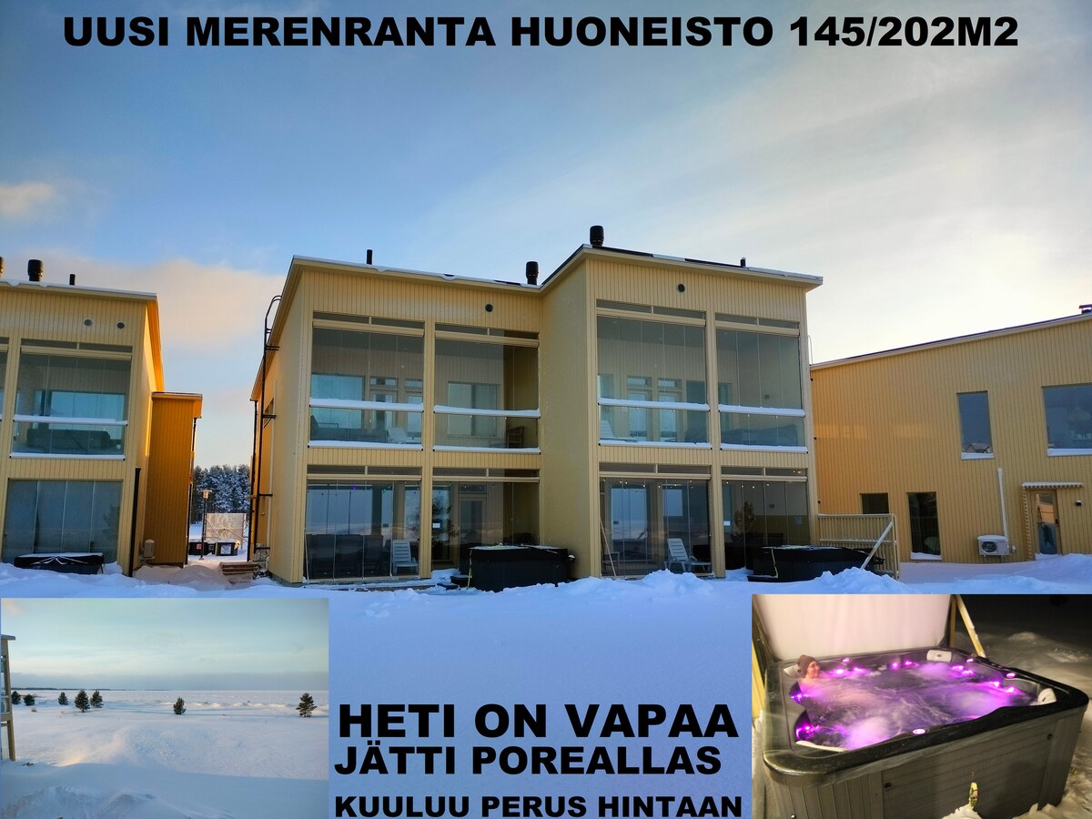 The bright yellow exterior of a two-story beachfront apartment complex is presented against a clear sky. Snow covers the ground, highlighting the structure's clean lines and large glass doors. A large hot tub with illuminated features is located in the foreground.